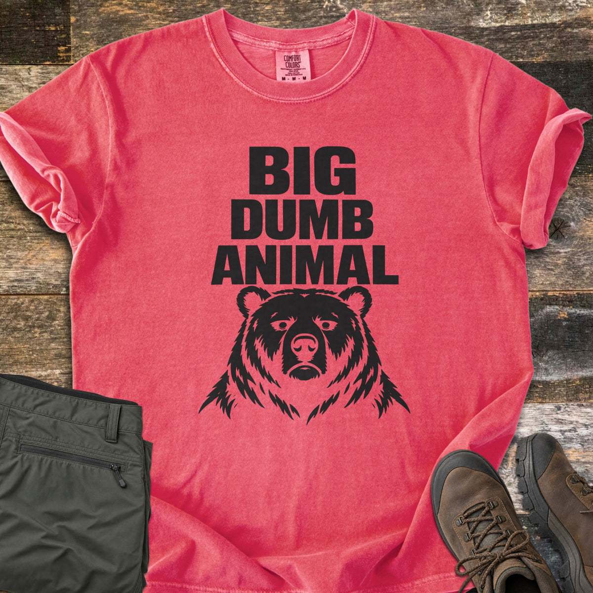 Big Dumb Animal Comfort Colors T-shirt in Watermelon, laid flat. Bold black block letters read Big Dumb Animal above a grumpy-faced bear head. The shirt, sleeves rolled, lies on weathered wood planks beside folded olive hiking pants and brown hiking boots.