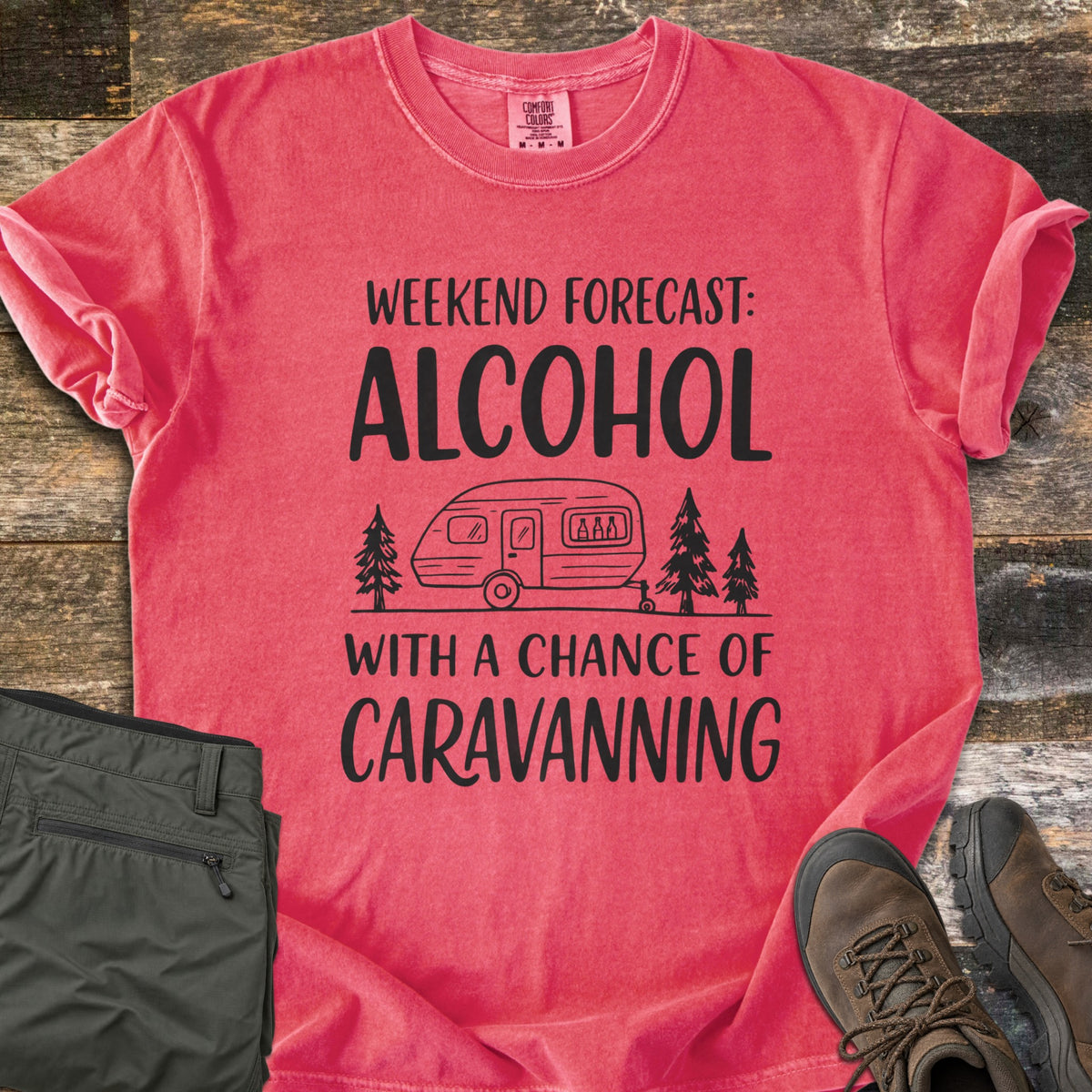 Alcohol With a Chance of Caravanning Comfort Colors T-shirt in Watermelon, laid flat. Black print reads Weekend Forecast: Alcohol above a caravan with bottles in its window between pine trees, then With a Chance of Caravanning. Olive trousers and brown hiking boots sit on dark wood planks.