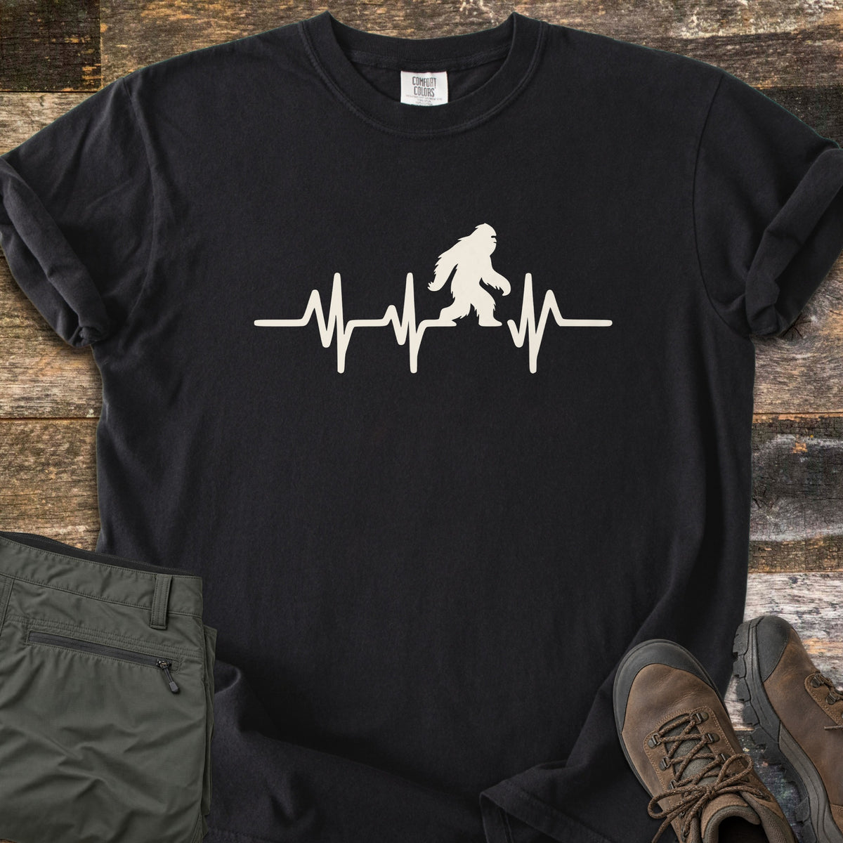 Bigfoot Heartbeat Comfort Colors T-shirt in Black, laid flat. A cream heartbeat line runs across the chest with a walking Bigfoot silhouette in the middle; there are no words. The shirt lies on weathered wood beside folded olive hiking pants and brown hiking boots.