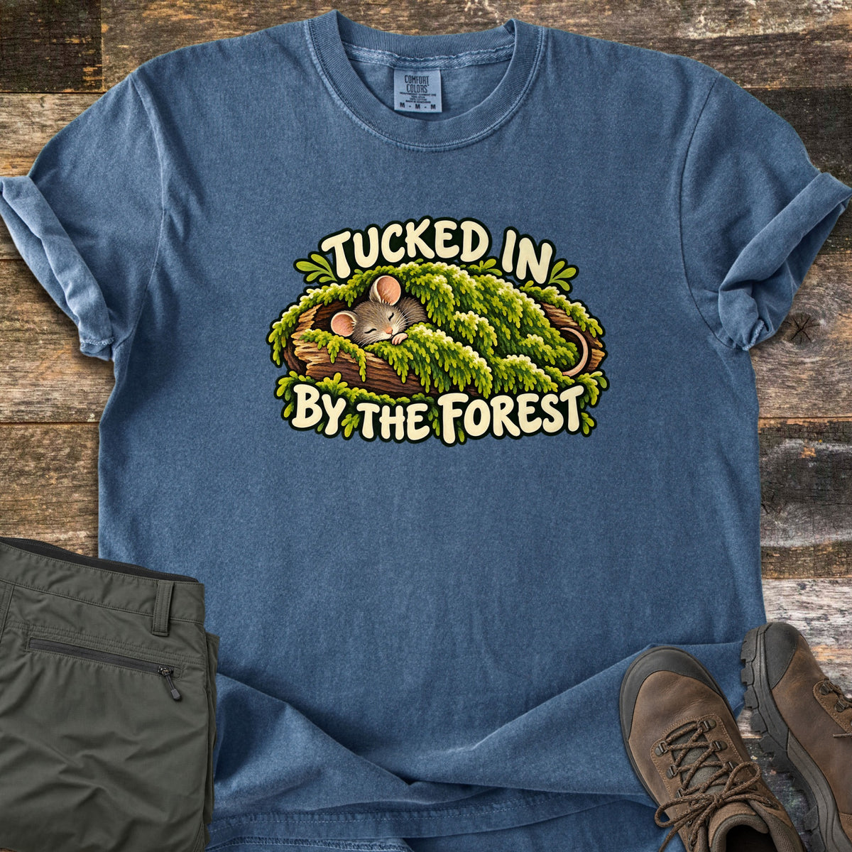 Tucked in by the Forest Comfort Colors T-shirt in Denim, laid flat. A sleepy mouse tucked under green moss on a log, framed by the words Tucked In By the Forest. Set on weathered wood planks with olive hiking pants and brown hiking boots beside it.