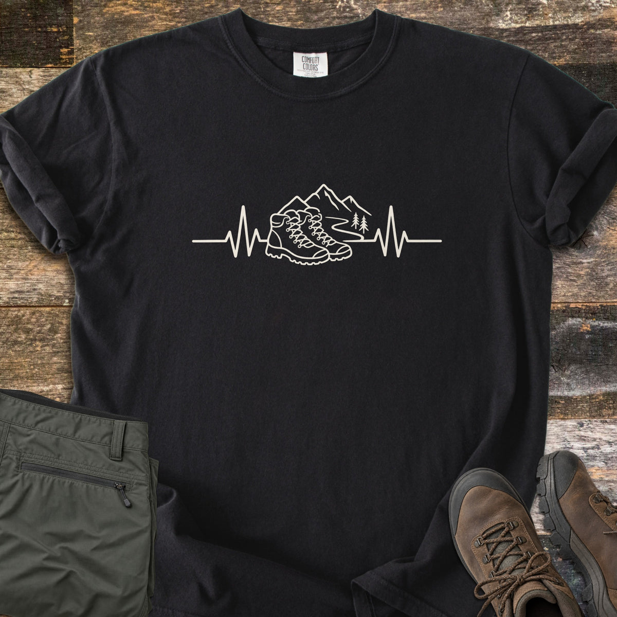 Hiking Heartbeat Comfort Colors T-shirt in Black, laid flat. A cream line drawing of hiking boots, mountains, a winding trail and two pine trees sits in a heartbeat line, with no words. Cuffed sleeves on weathered wood beside folded olive pants and brown boots.