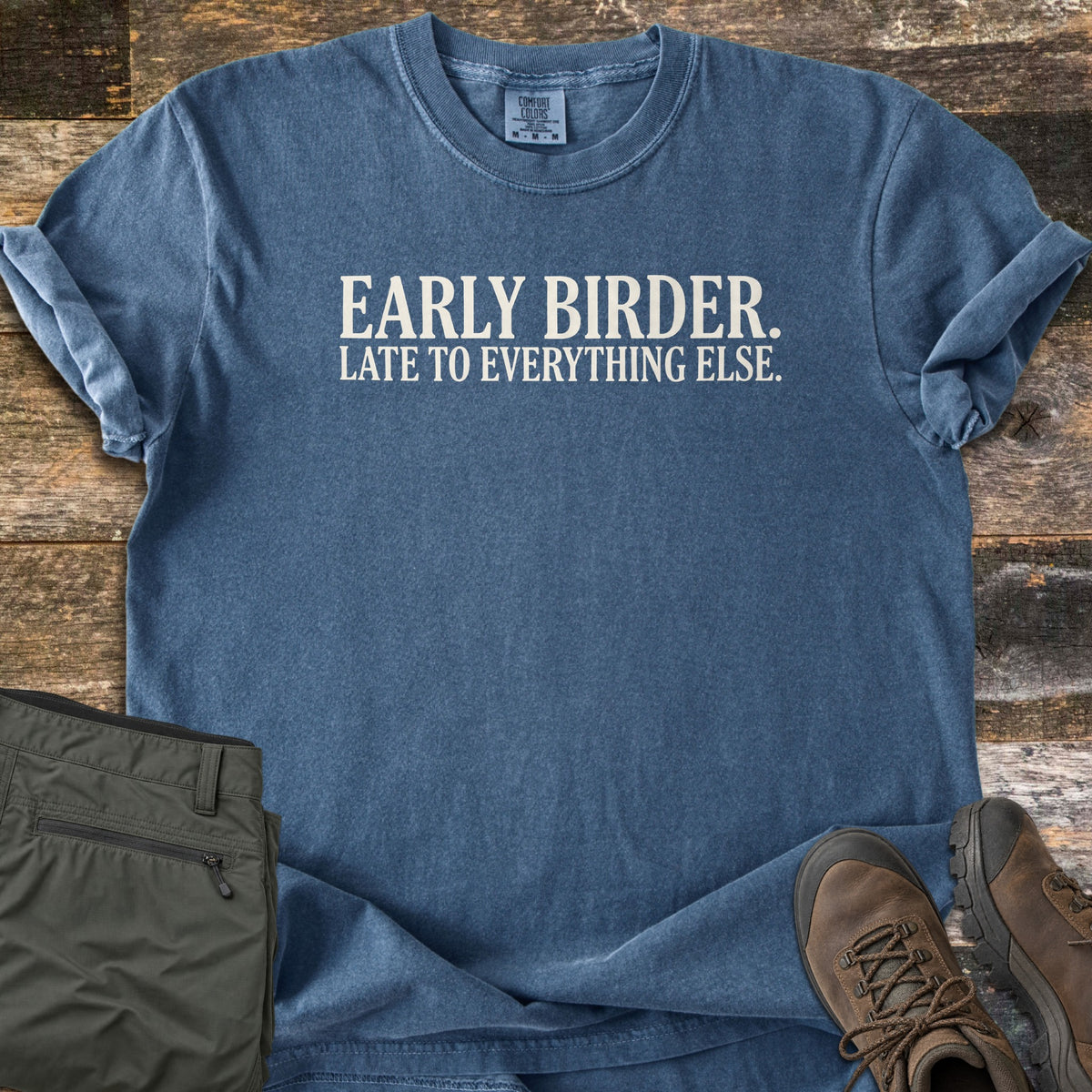Early Birder Comfort Colors T-shirt in Denim, laid flat. Cream serif text across the chest reads EARLY BIRDER. LATE TO EVERYTHING ELSE. The shirt lies on weathered wood planks beside folded olive hiking pants and a pair of brown leather hiking boots.