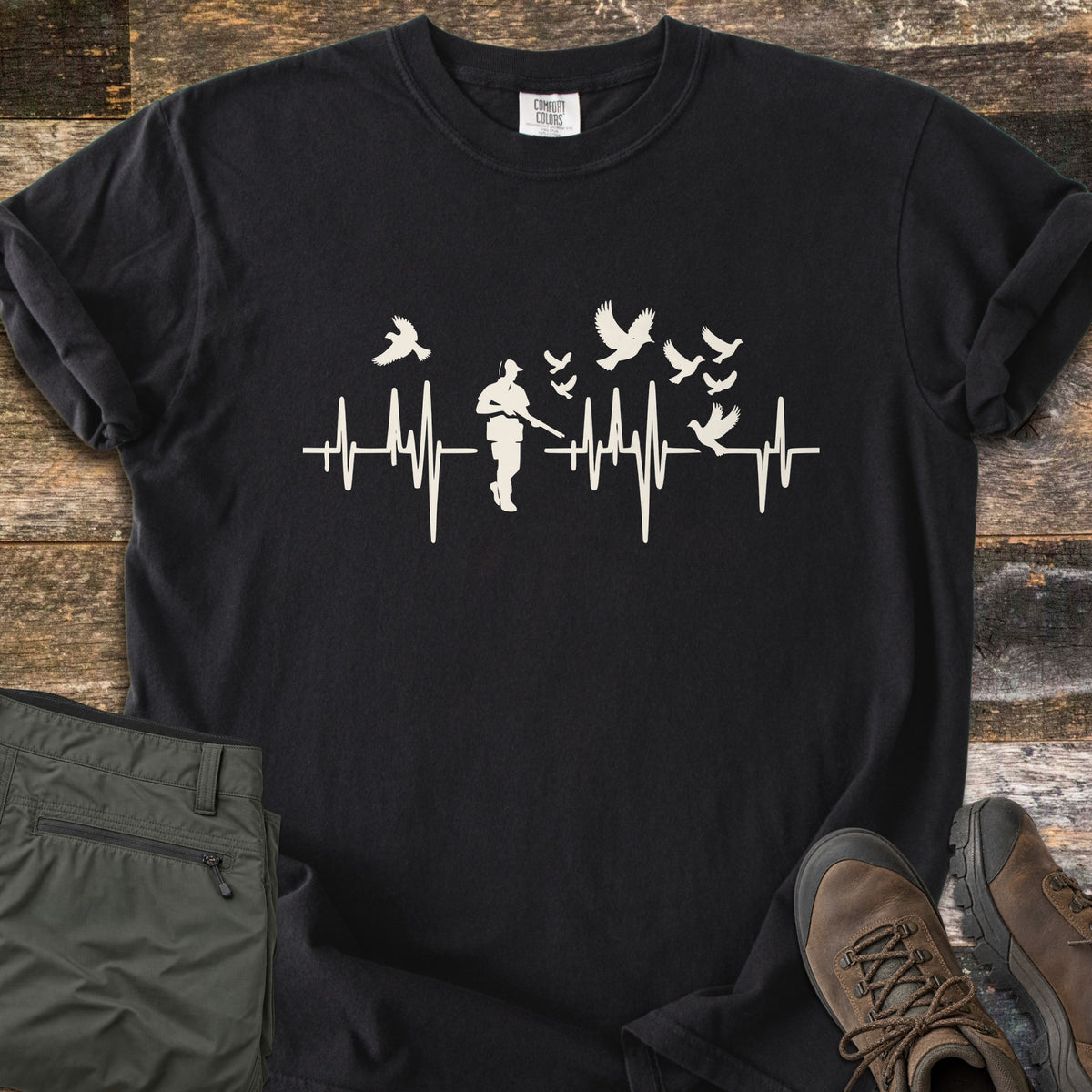 Bird Hunter Comfort Colors T-shirt in Black, laid flat. A cream heartbeat line crosses the chest with a hunter silhouette in a cap holding a shotgun and birds flying up from the line. Folded olive pants and brown hiking boots lie beside it on weathered wood.