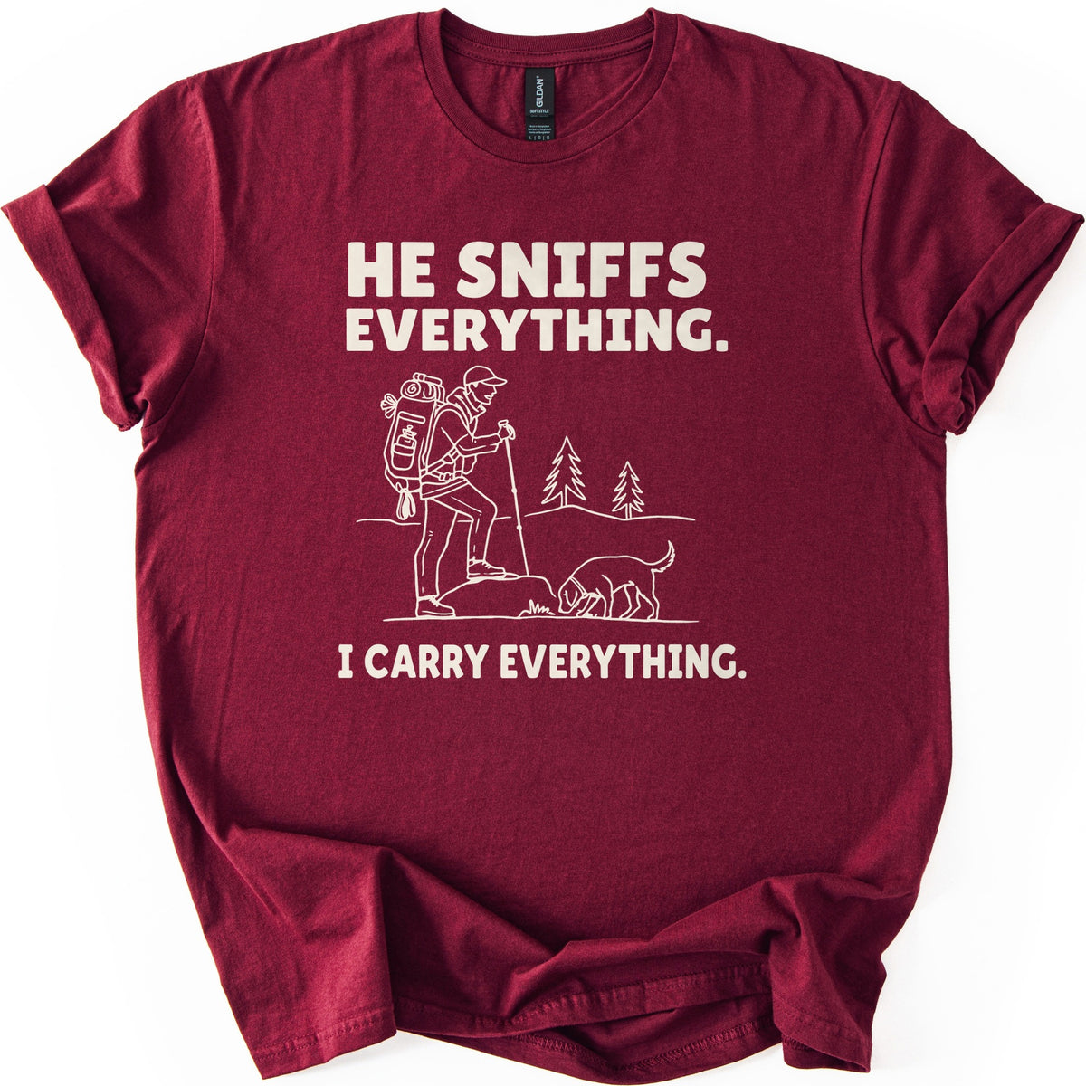 Trail Dog Pack Mule Comfort Colors T-shirt in Maroon, laid flat. Cream line art of a hiker with a loaded backpack and trekking pole stepping onto a rock as a dog sniffs the ground, with pine trees behind, between the words He sniffs everything. and I carry everything. Sleeves rolled, on white.
