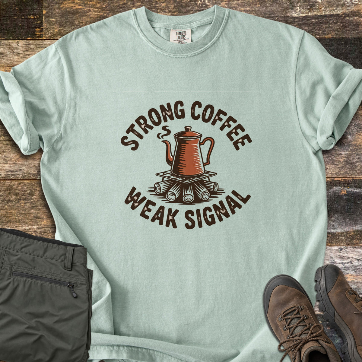 Strong Coffee Weak Signal Comfort Colors T-shirt in Bay, laid flat. A steaming copper coffee pot sits on a grate over campfire logs, arched by the words Strong Coffee above and Weak Signal below. Folded olive hiking pants and brown hiking boots lie on weathered wood planks.