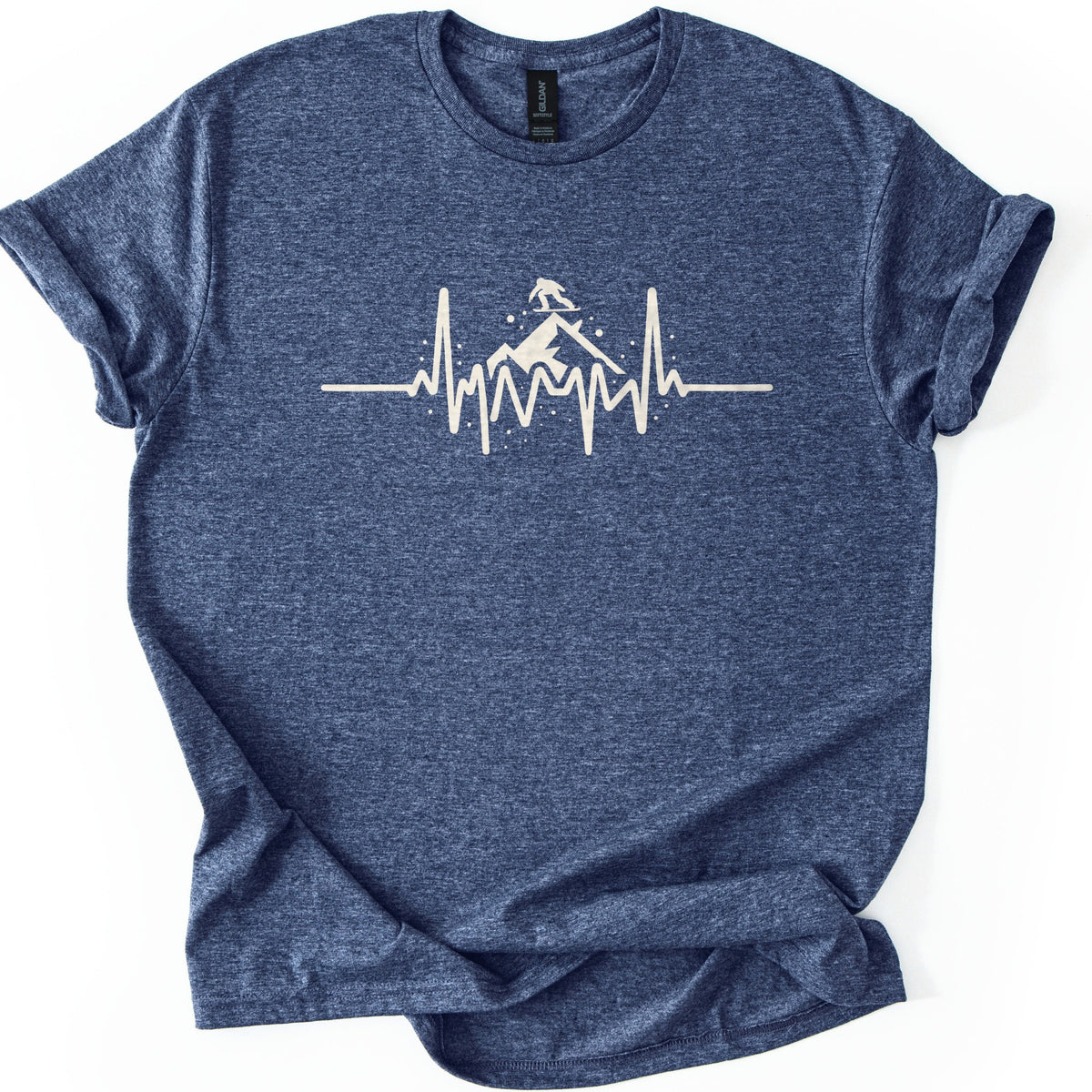 Snow Boarder Heartbeat Comfort Colors T-shirt in Heather Navy, laid flat. A cream heartbeat line rises into a snowy mountain peak with a snowboarder silhouette riding on top, surrounded by snow dots. No words. The heathered shirt has rolled sleeves and lies on a white background.