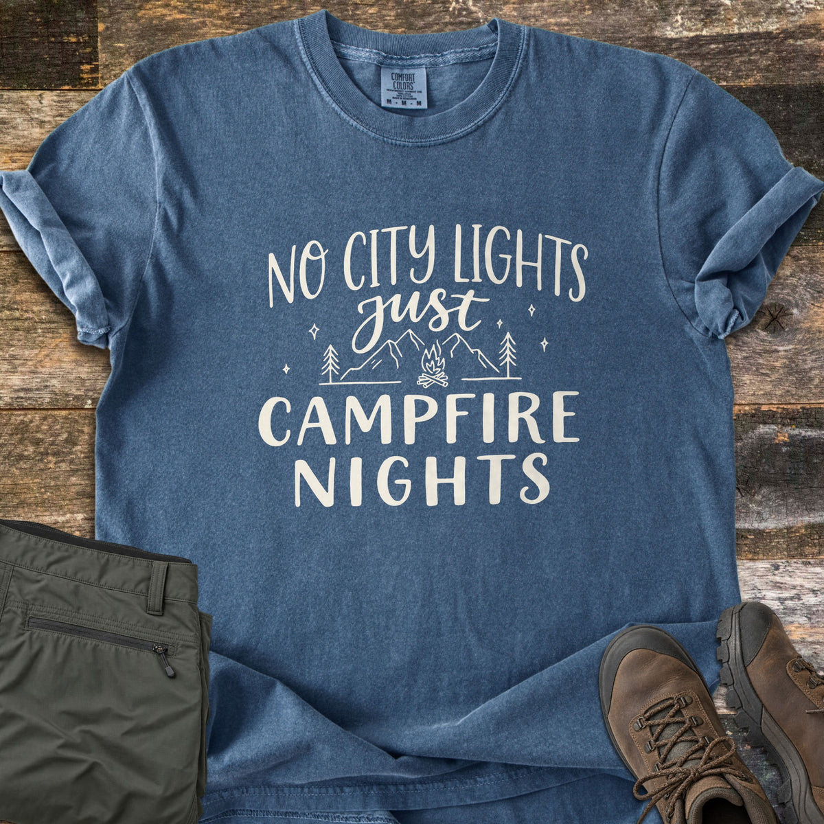 Campfire Nights Comfort Colors T-shirt in Denim, laid flat. White text reads No City Lights Just Campfire Nights around line art of mountains, pine trees, stars and a campfire. It lies on weathered wood beside olive hiking pants and brown hiking boots.