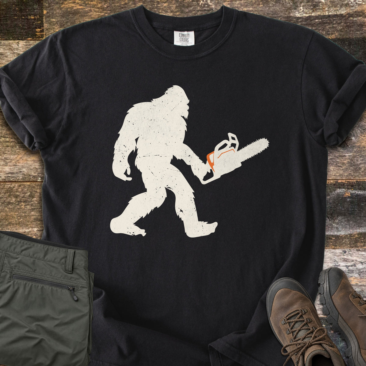 Lumber Squatch Comfort Colors T-shirt in Black, laid flat. A distressed cream silhouette of Bigfoot walks while carrying a chainsaw with orange accents. The sleeves are rolled, with folded olive pants and brown hiking boots on rustic wood planks.