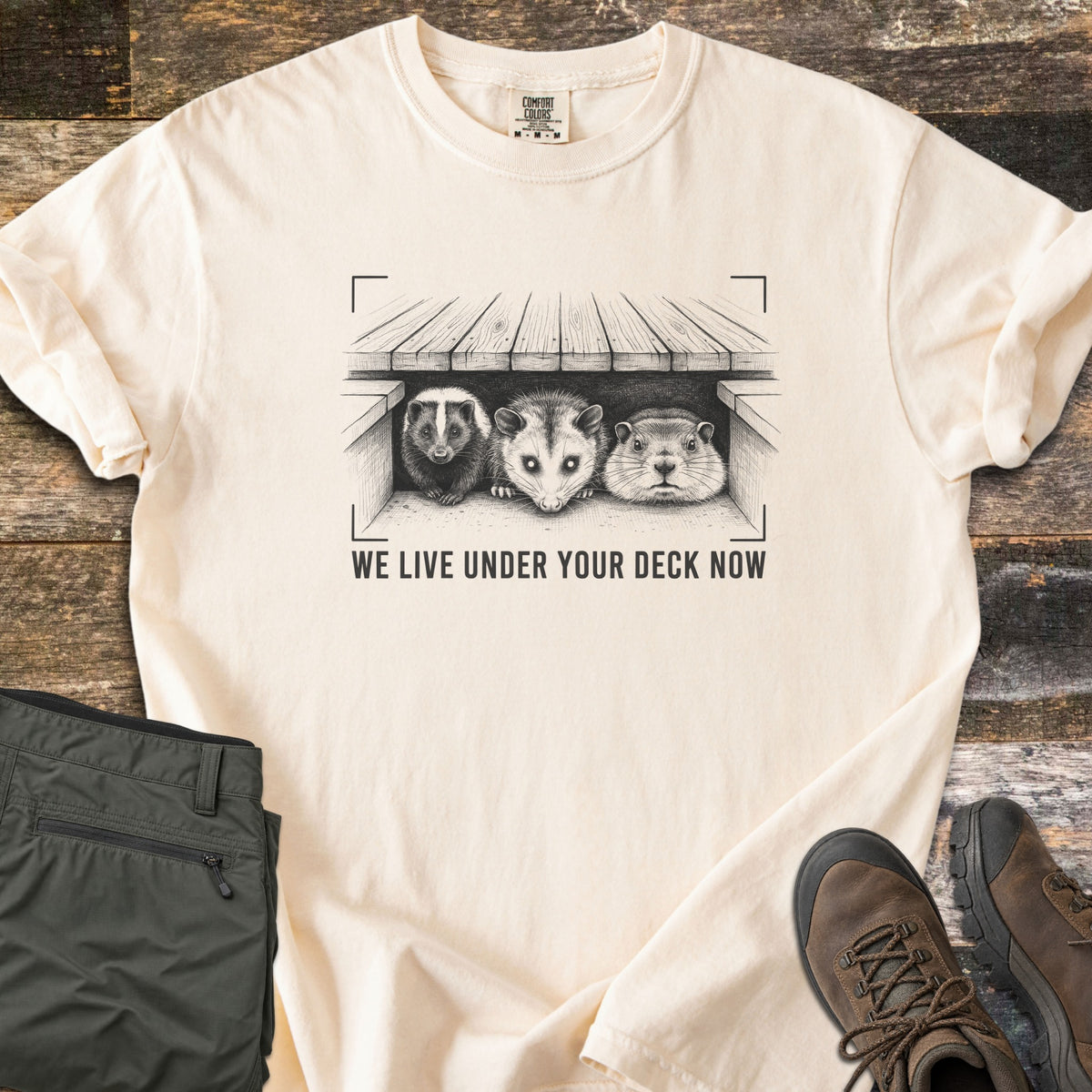 We Live Under Your Deck Now Comfort Colors T-shirt in Ivory, laid flat. In dark ink, a sketched skunk, opossum and groundhog peer from under a wooden deck inside viewfinder corners, above the words We Live Under Your Deck Now. Olive pants and brown hiking boots lie on weathered boards.