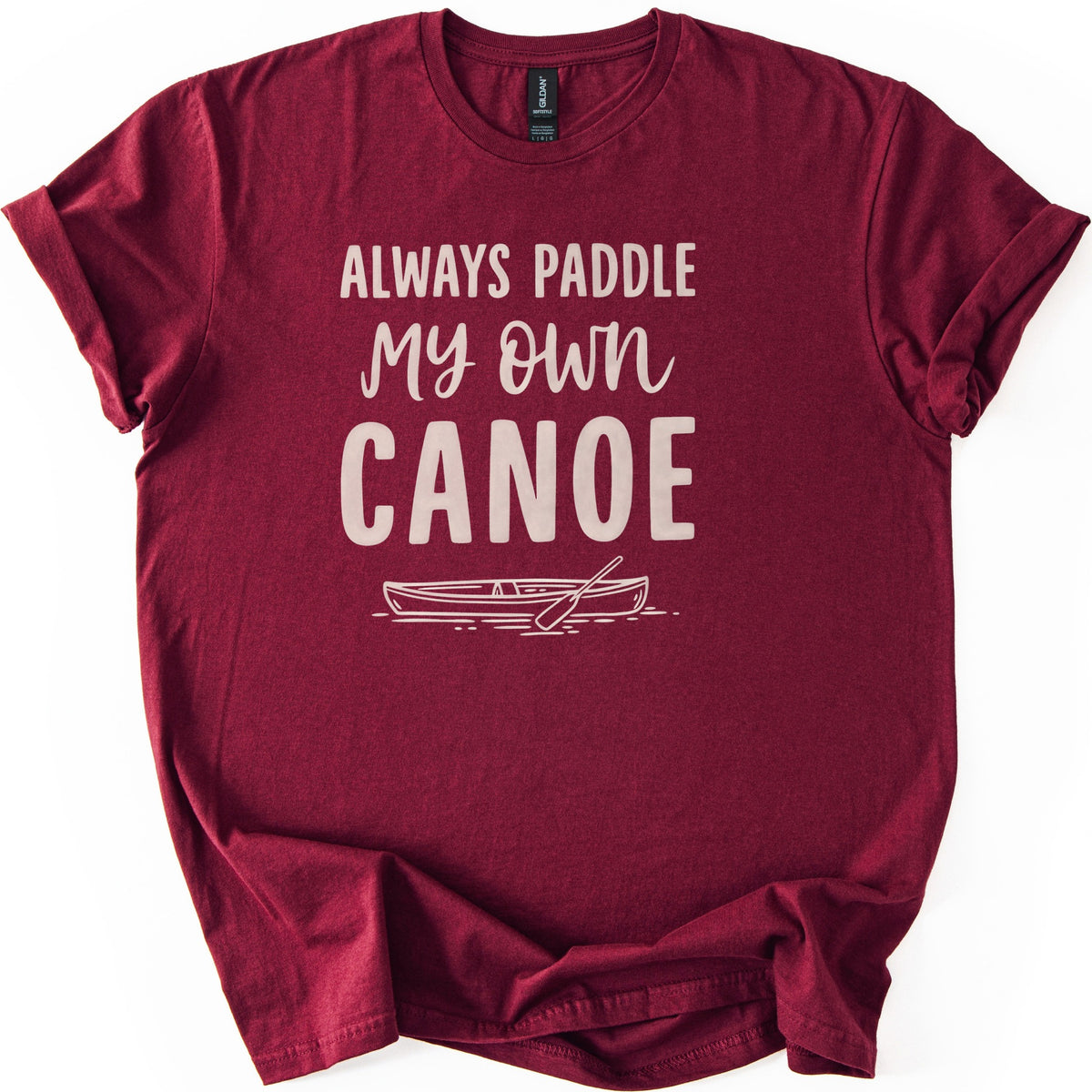 Paddle My Own Canoe Comfort Colors T-shirt in Maroon, laid flat. Light pink text reads Always Paddle My Own Canoe above a line drawing of a canoe with a paddle on rippling water. The shirt has rolled sleeves and lies on a white surface.