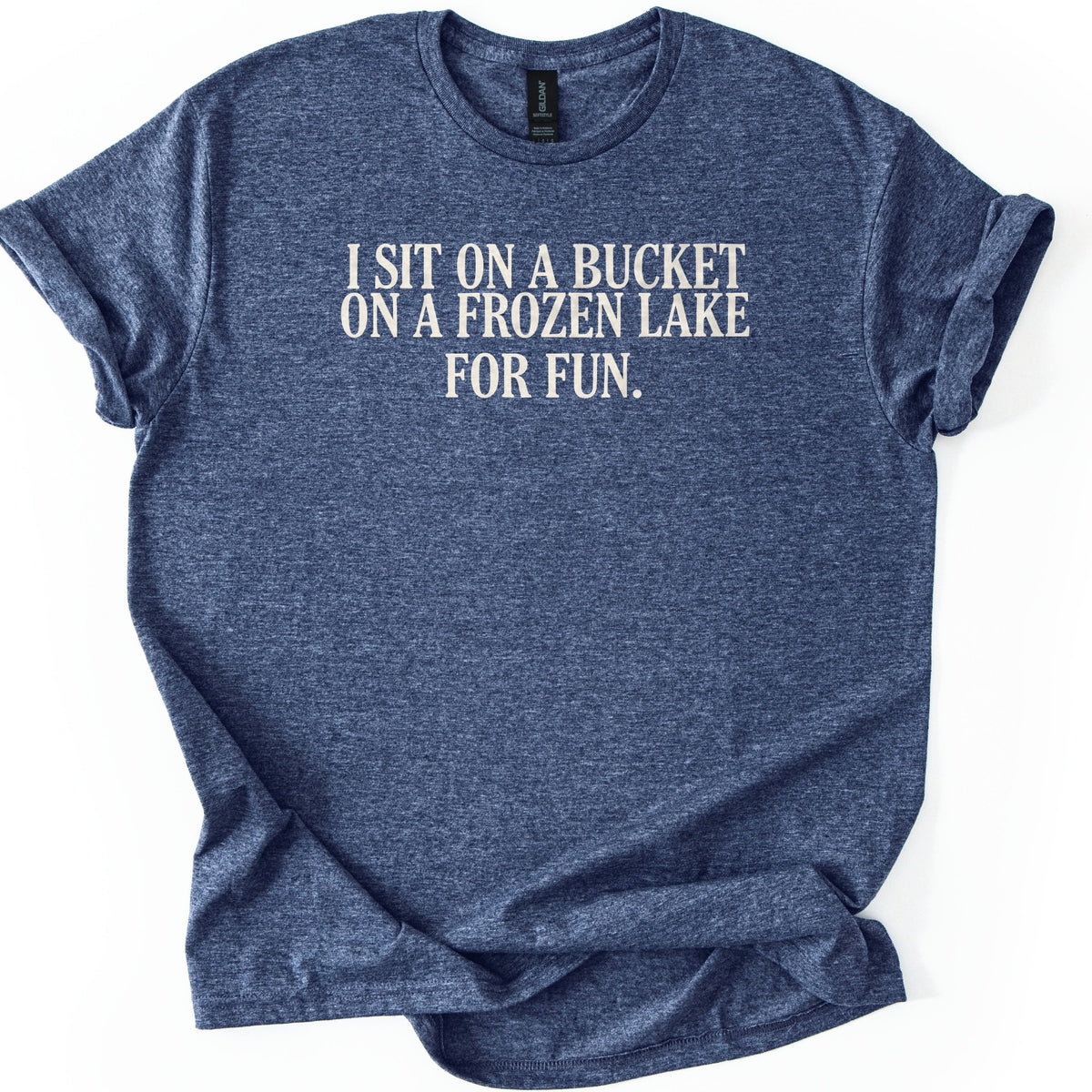Bucket On A Frozen Lake Comfort Colors T-shirt in Heather Navy, laid flat. Cream serif capital letters across the chest read I SIT ON A BUCKET ON A FROZEN LAKE FOR FUN. The shirt lies on a white surface with its sleeves rolled and the hem loosely folded.