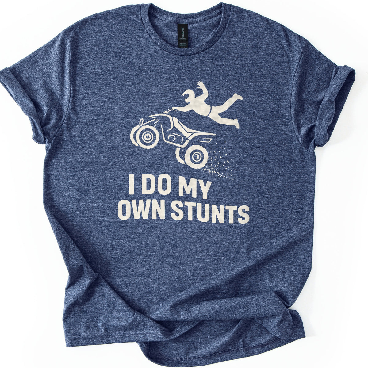 ATV Stunts Comfort Colors T-shirt in Heather Navy, laid flat. A cream silhouette of a helmeted rider flying off the back of an ATV as it kicks up dirt, above the words I DO MY OWN STUNTS. The shirt lies on a white background with its sleeves cuffed.