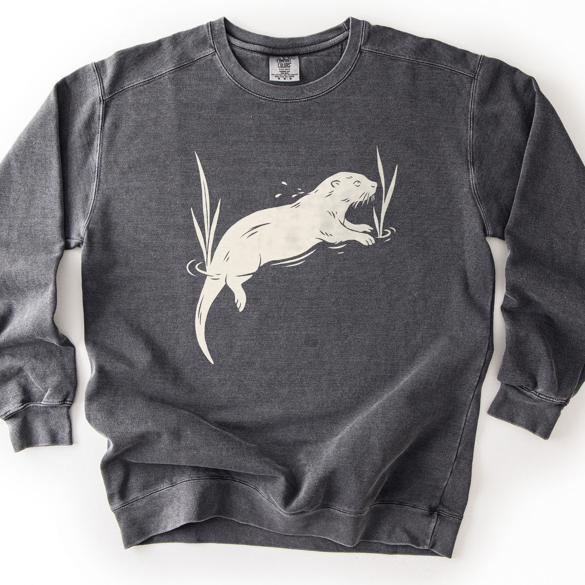 Swimming Otter Comfort Colors crewneck sweatshirt in Pepper, laid flat. A cream block-print style otter swims with its head raised, splashing droplets, between two tall reeds with ripples around them. No words are printed. The sweatshirt lies on a white background.