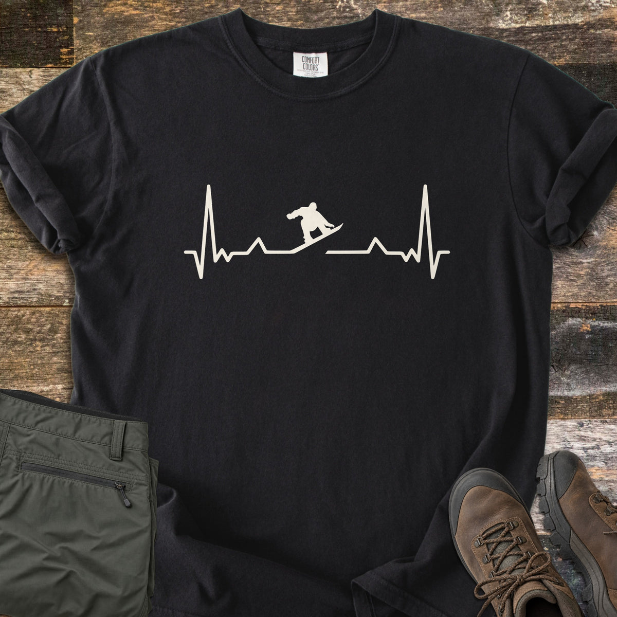 Snowboarder Heartbeat Comfort Colors T-shirt in Black, laid flat. A cream heartbeat line runs across the chest, with a snowboarder silhouette riding the line in the middle. Sleeves are rolled, on a rustic wood floor beside folded olive pants and brown hiking boots.