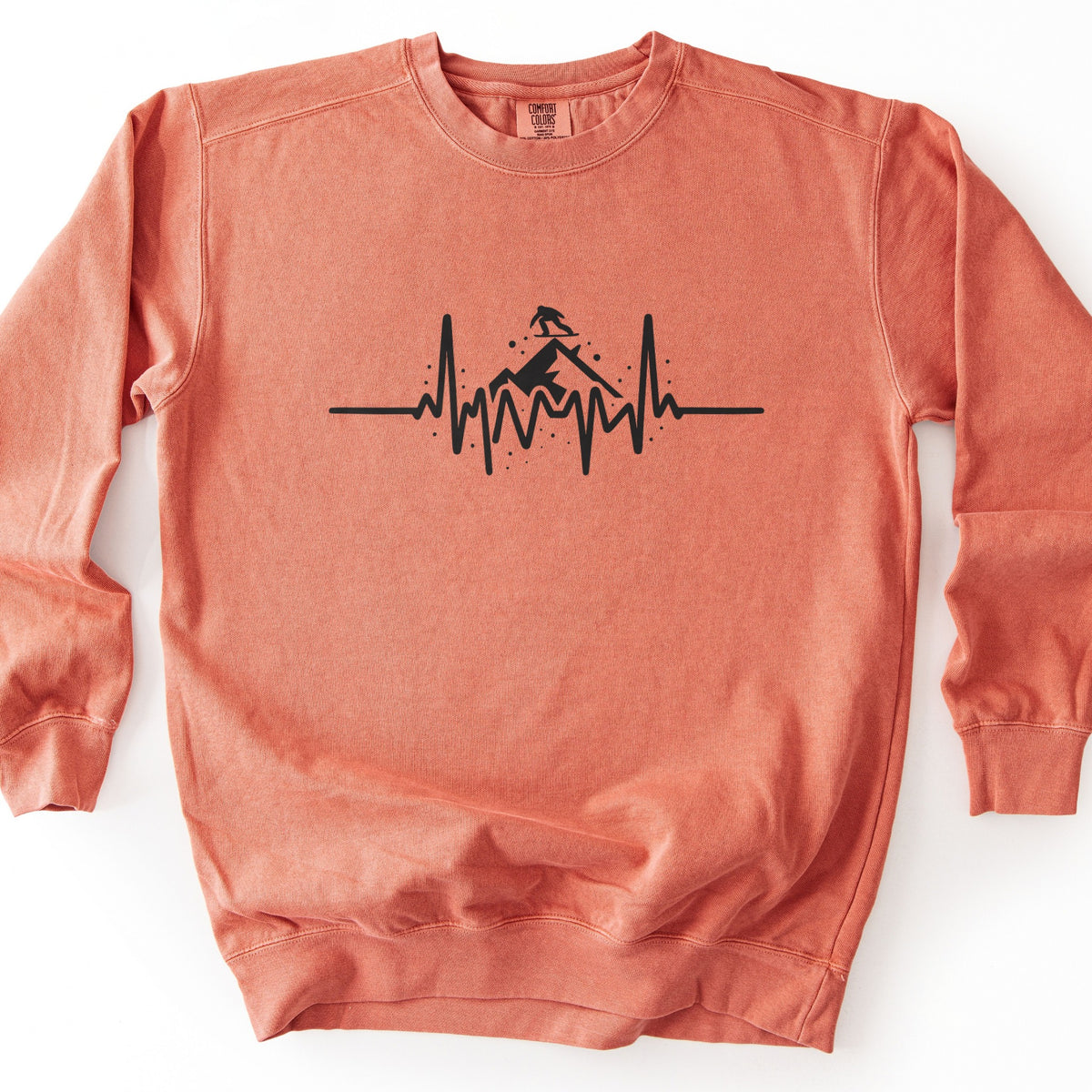 Snow Boarder Heartbeat Comfort Colors crewneck sweatshirt in Terracotta, laid flat. A black heartbeat line runs across the chest, rising into a mountain peak with a snowboarder riding on top and small dots of snow around it. No words. The sweatshirt lies on a white surface.