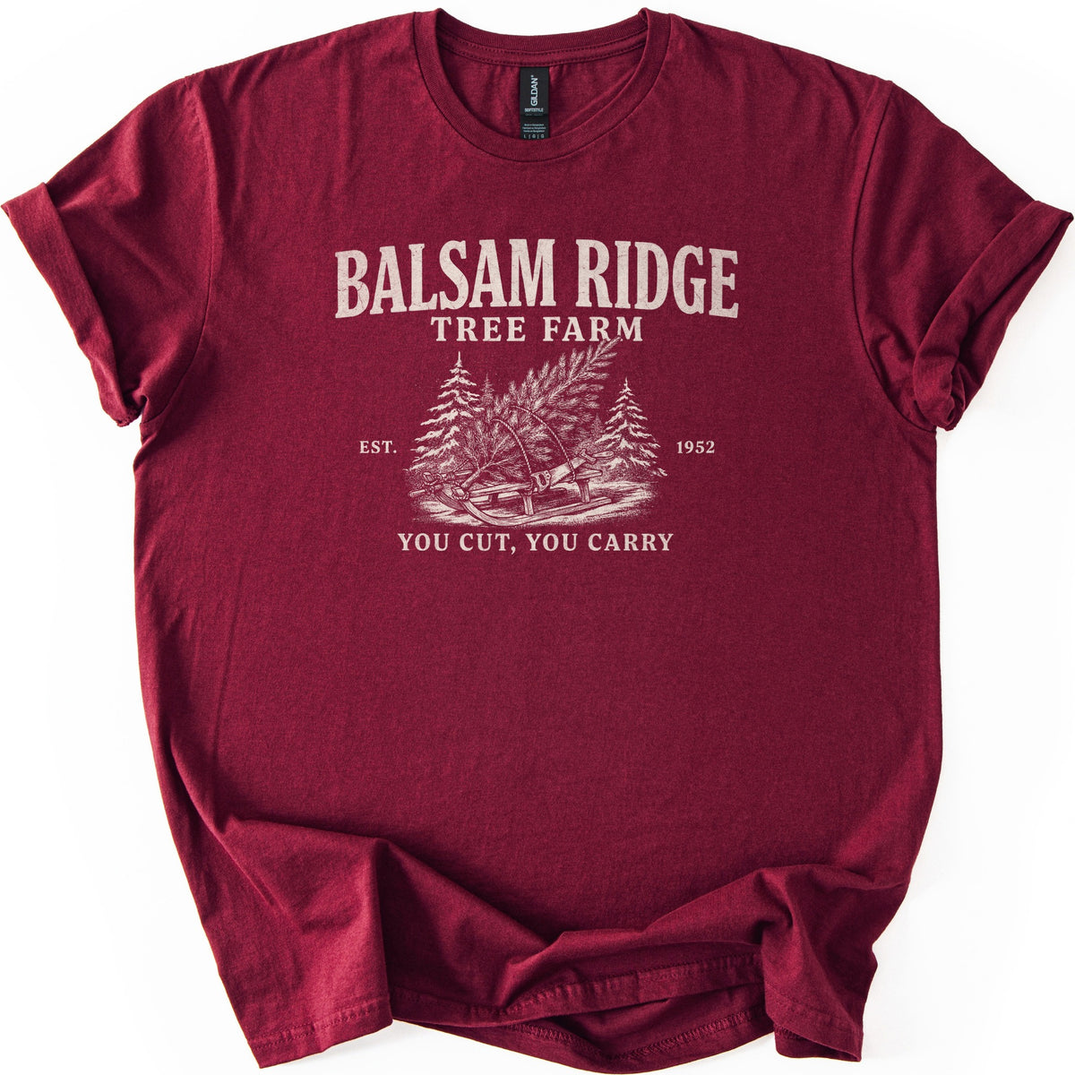 Balsam Ridge Tree Farm Comfort Colors T-shirt in Maroon, laid flat. Off-white print reads Balsam Ridge Tree Farm above an engraved-style fir tree tied to a wooden sled between snowy pines, flanked by Est. 1952, with You Cut, You Carry below. Sleeves cuffed on a white surface.
