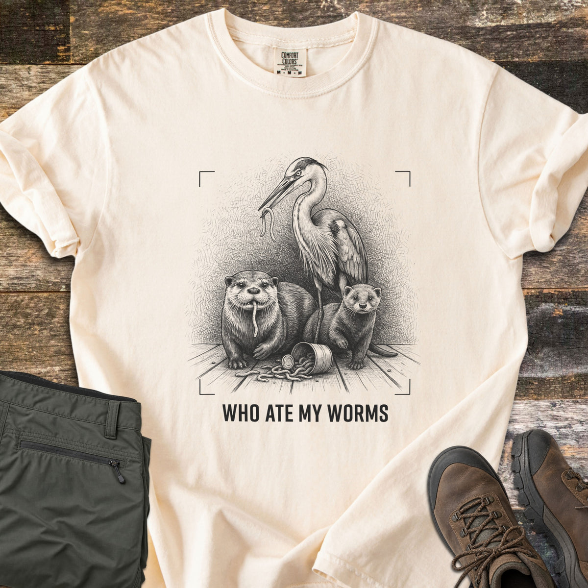 Who Ate My Worms Comfort Colors T-shirt in Ivory, laid flat. A black sketch of a heron and an otter each eating a worm beside a mink and a spilled bait can, inside viewfinder corners, above the words Who Ate My Worms. Olive pants and brown hiking boots lie on weathered wood.