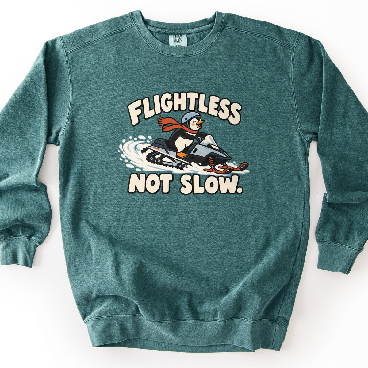 Flightless Not Slow Comfort Colors crewneck sweatshirt in Blue Spruce, laid flat. A cartoon penguin in a gray helmet and red scarf rides a snowmobile through a spray of snow between the words Flightless and Not Slow. in cream lettering. Shown on a white background.