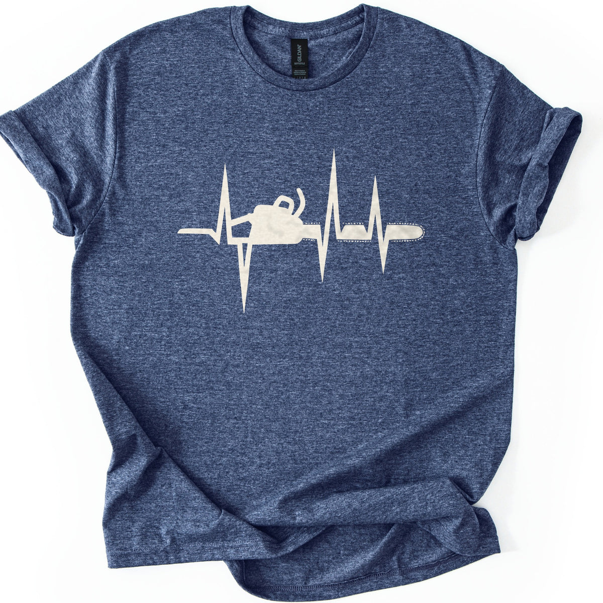 Chainsaw Heartbeat Comfort Colors T-shirt in Heather Navy, laid flat. A cream heartbeat line with sharp peaks runs across the chest, with a chainsaw silhouette set into the line, its bar and chain extending to the right. The sleeves are rolled on a white background.