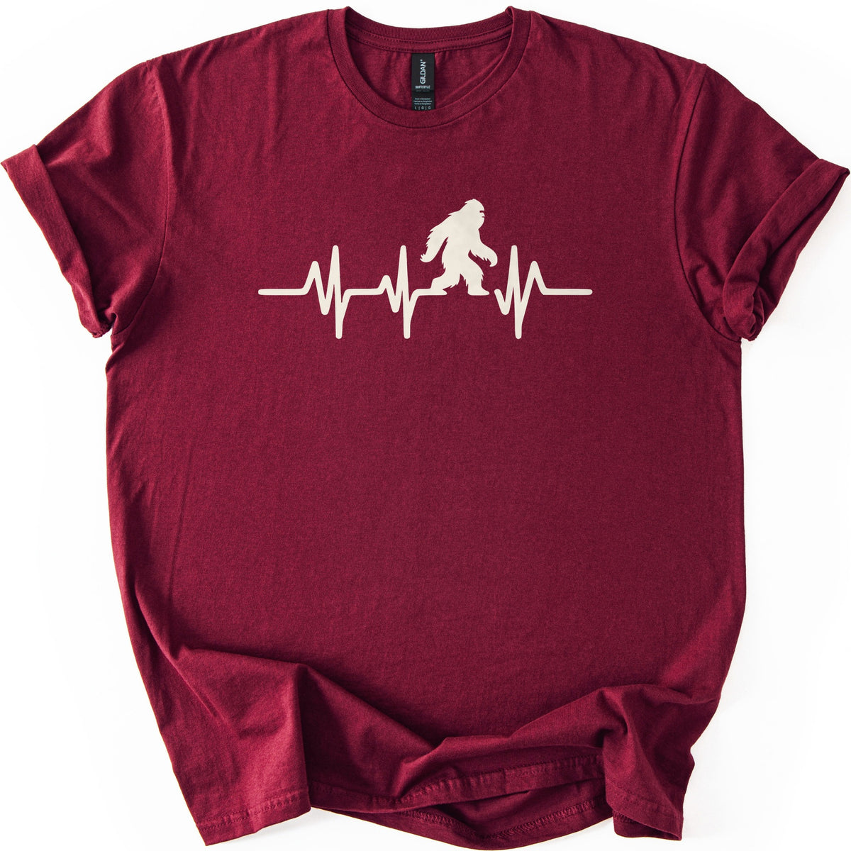Yeti Heartbeat Comfort Colors T-shirt in Maroon, laid flat. A cream-colored heartbeat line runs across the chest, with a walking Bigfoot silhouette standing within it; there are no words. The sleeves are cuffed and the shirt lies on a white background.