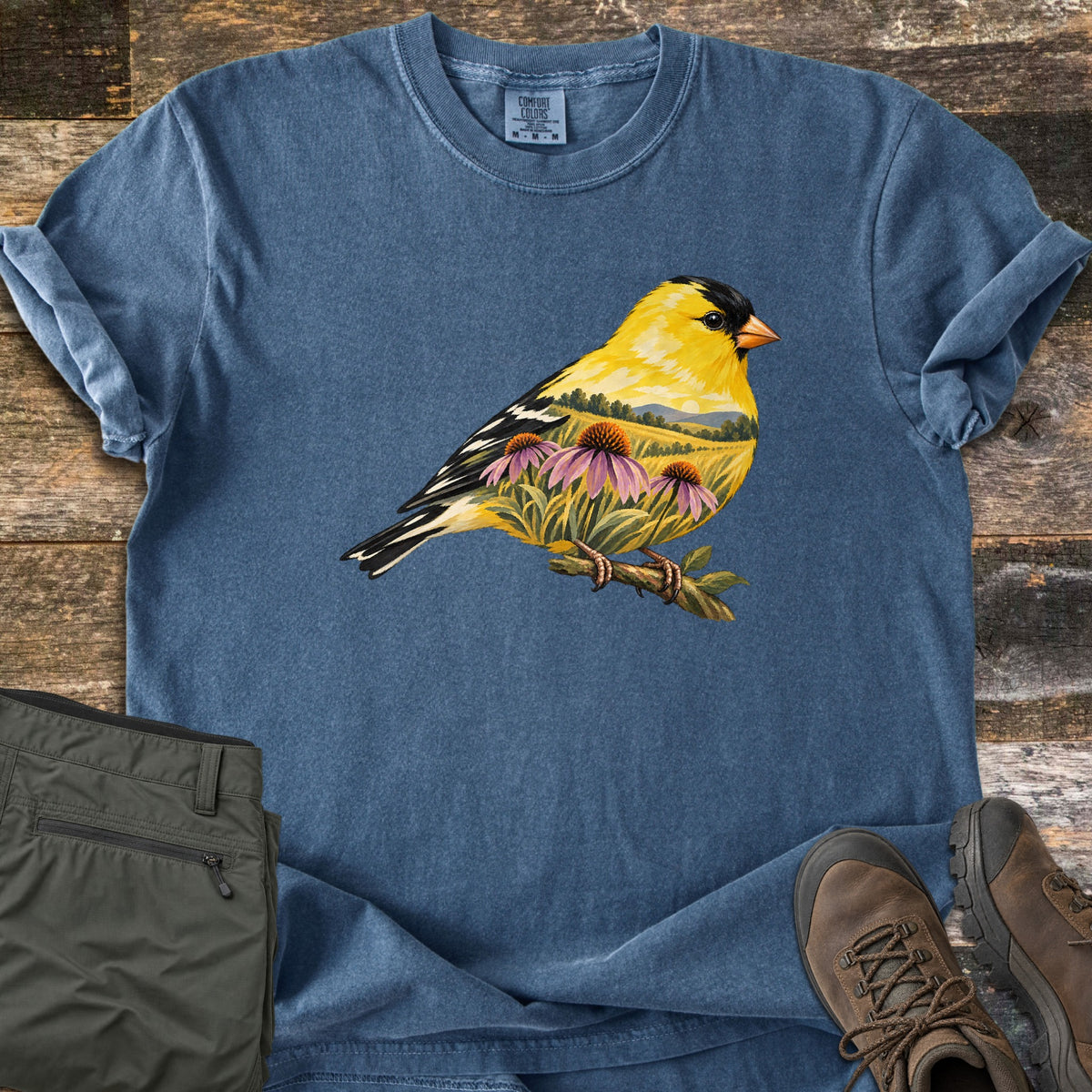 Goldfinch Meadow Comfort Colors T-shirt in Denim, laid flat. A yellow and black goldfinch perches on a twig, its body filled with a meadow scene of rolling hills and pink coneflowers. The shirt lies on weathered wood beside folded olive pants and brown hiking boots.
