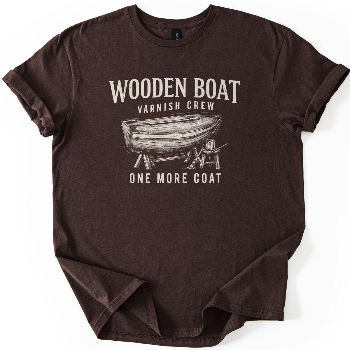 Wooden Boat Varnish Crew Comfort Colors T-shirt in Dark Chocolate, laid flat. Cream print reads Wooden Boat, Varnish Crew above a sketch of a wooden rowboat on sawhorses beside a stool with a varnish can and brush, then One More Coat. Sleeves cuffed on a white background.