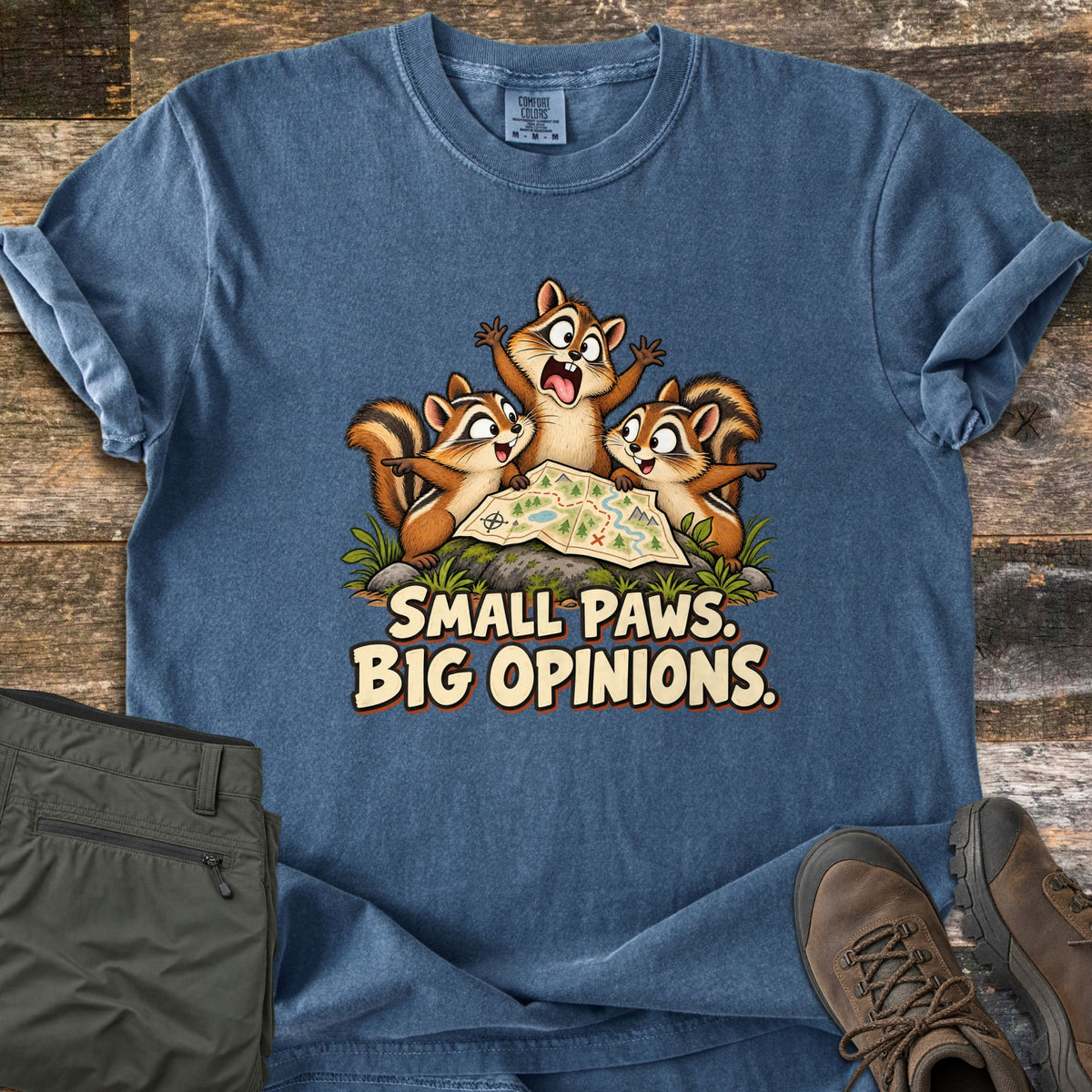 Small Paws. Big Opinions. Comfort Colors T-shirt in Denim, laid flat. Three cartoon chipmunks argue and point over a trail map on a mossy rock above the words Small Paws. Big Opinions. Olive hiking pants and brown hiking boots lie beside it on weathered wood planks.