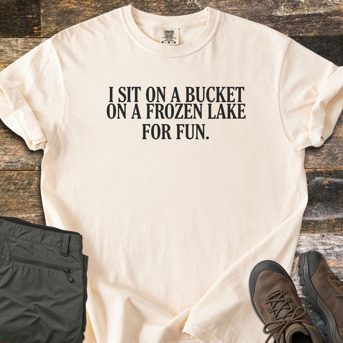 Bucket On A Frozen Lake Comfort Colors T-shirt in Ivory, laid flat. Black serif text reads I SIT ON A BUCKET ON A FROZEN LAKE FOR FUN. The shirt has rolled sleeves and lies on weathered wood planks beside folded olive hiking pants and brown hiking boots.