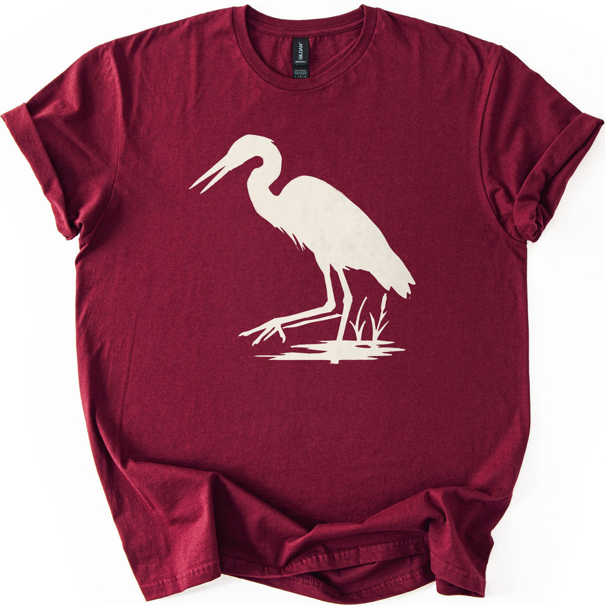 Great Blue Heron Comfort Colors T-shirt in Maroon, laid flat. A cream silhouette of a heron wading in shallow water beside cattails, beak open and one foot reaching forward. The shirt lies on a white surface with cuffed sleeves.