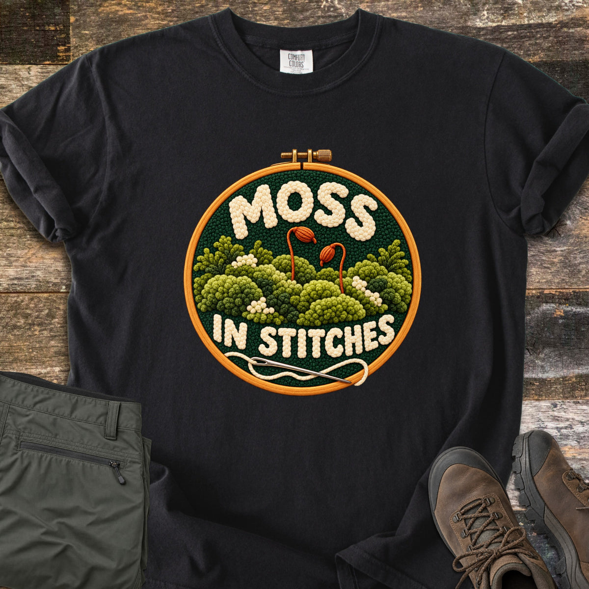 Moss in Stitches Comfort Colors T-shirt in Black, laid flat. An embroidery hoop holds stitched green moss with two orange spore capsules, the word Moss above and In Stitches below, plus a needle and thread. Folded olive pants and brown hiking boots lie beside it on weathered wood.