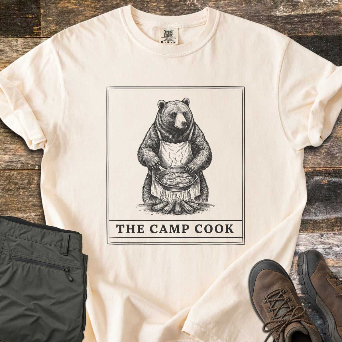 The Camp Cook Comfort Colors T-shirt in Ivory, laid flat. A framed sketch of a bear in an apron frying bacon in a skillet over a campfire, above the words THE CAMP COOK. Sleeves are cuffed on weathered wood beside olive hiking pants and brown hiking boots.