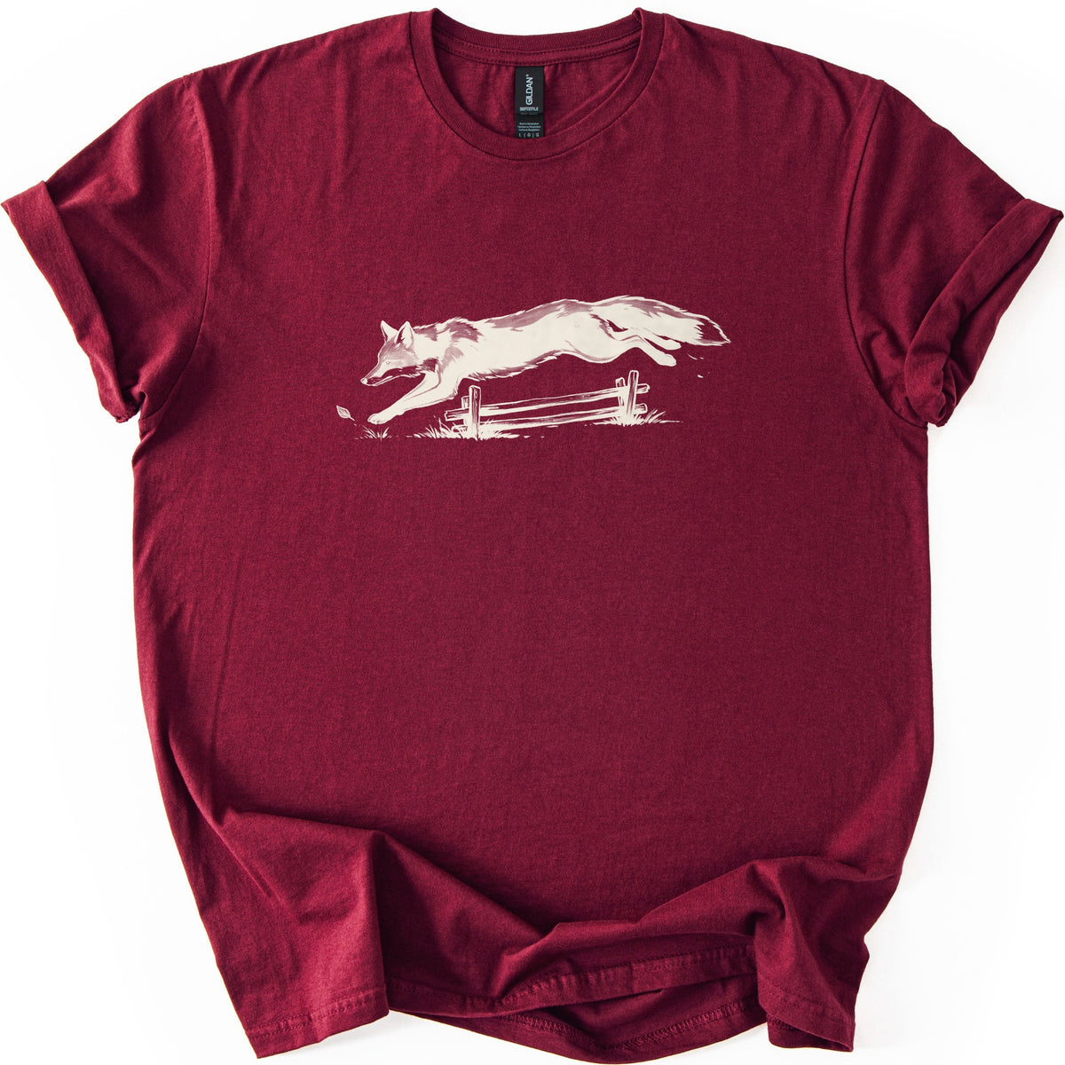 Leaping Fox Comfort Colors T-shirt in Maroon, laid flat. A cream-colored sketch of a fox leaping over a low wooden rail fence, with grass tufts and a falling leaf below. The shirt has cuffed sleeves and lies on a white background.