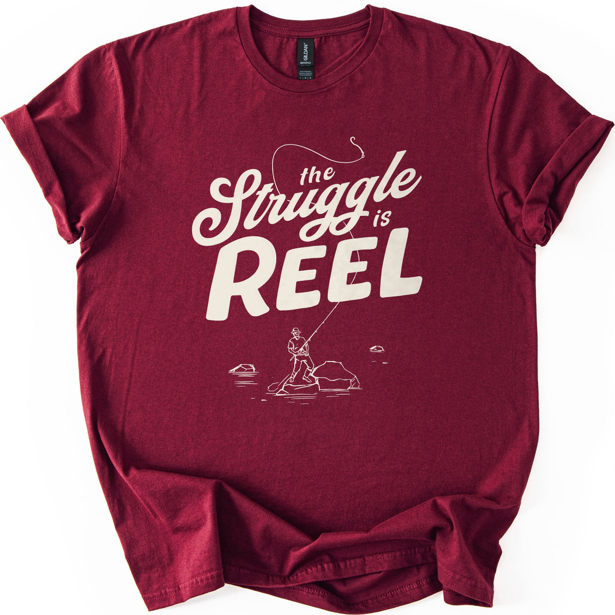 Struggle Is Reel Angler Comfort Colors T-shirt in Maroon, laid flat. Cream text reads the Struggle is REEL, with a fishing line arcing over the words to a hook. Below, a line drawing of an angler in a hat casts from a rock in the water. Sleeves cuffed, on a white background.