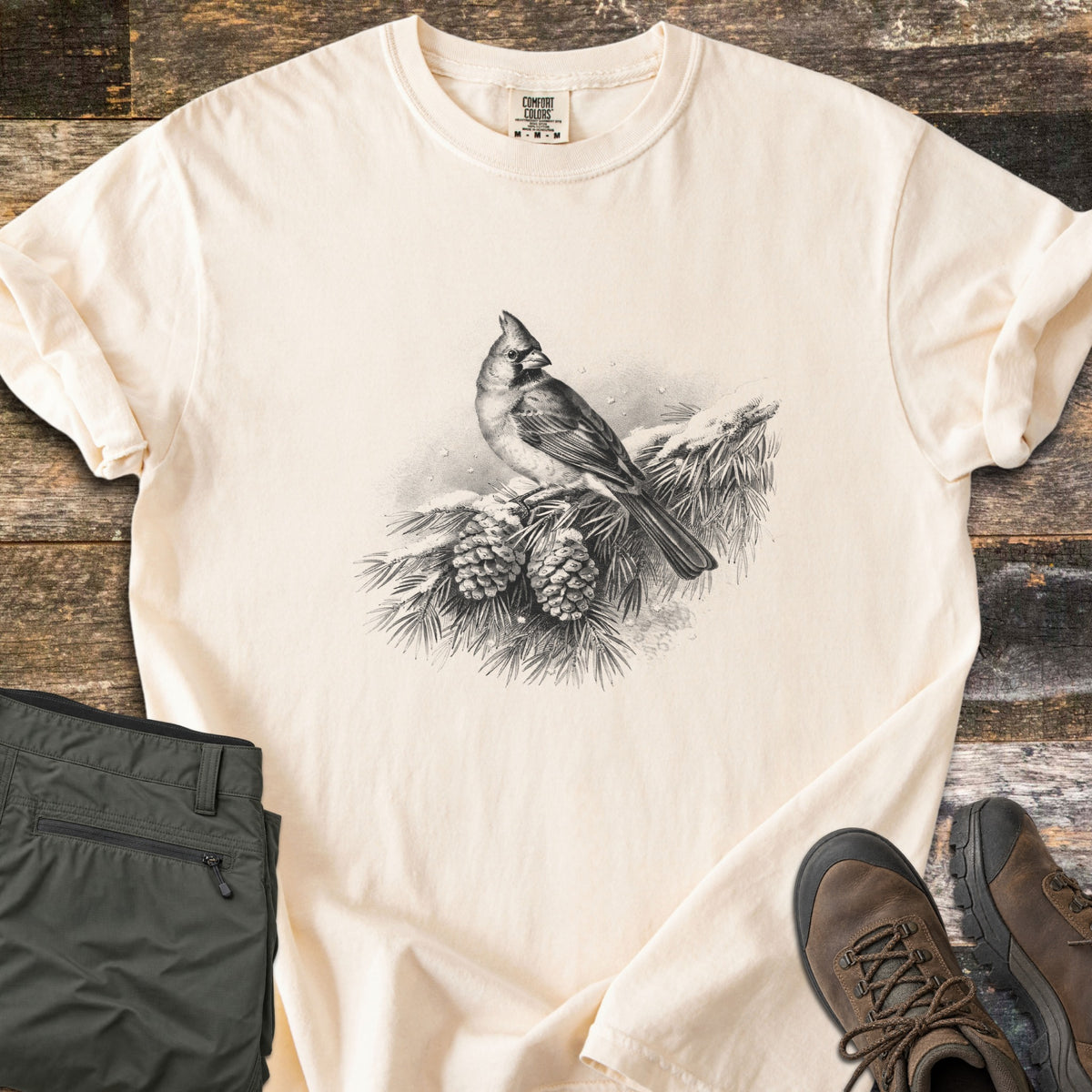 Cardinal On A Snowy Bough Comfort Colors T-shirt in Ivory, laid flat. A grayscale drawing of a cardinal perched on a snow-covered pine branch with two pinecones, with no words. Sleeves rolled, on weathered wood planks beside olive hiking pants and brown hiking boots.