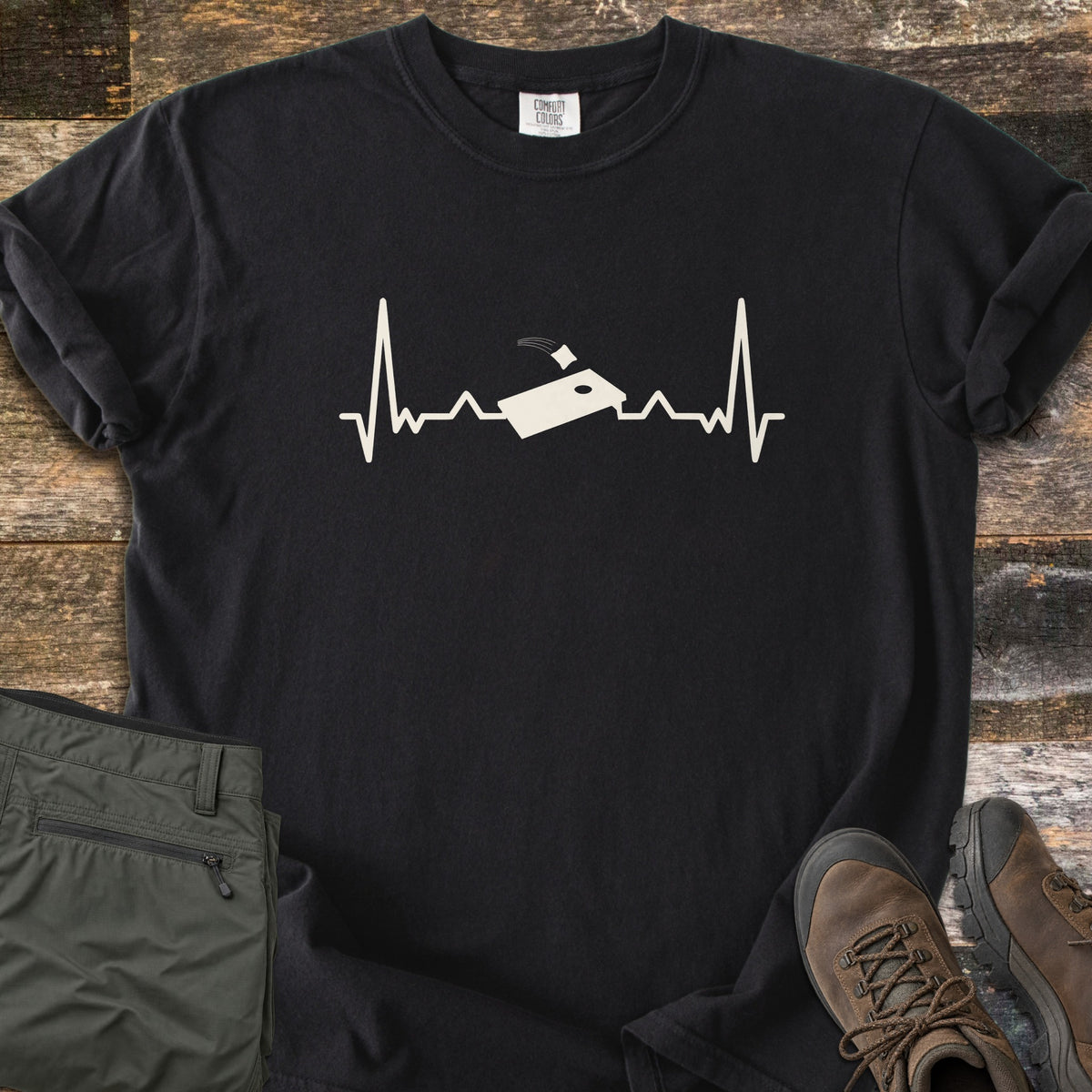 Corn Hole Comfort Colors T-shirt in Black, laid flat. A cream heartbeat line runs across the chest with a cornhole board at its center and a bean bag flying toward the hole. Olive pants and brown hiking boots sit beside it on weathered wood planks.