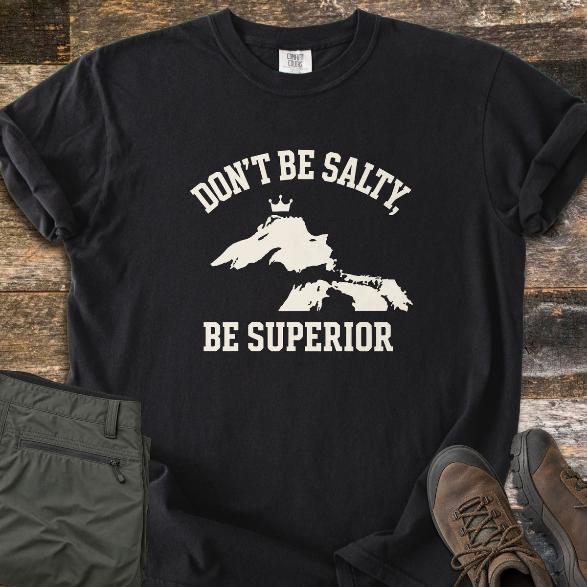 Be Superior Comfort Colors T-shirt in Black, laid flat. Cream text reads Don't Be Salty, arched over a Lake Superior silhouette topped with a small crown, with Be Superior below. Sleeves rolled, on weathered wood with olive hiking pants and brown hiking boots.