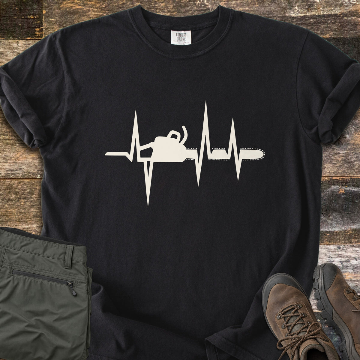 Chainsaw Heartbeat Comfort Colors T-shirt in Black, laid flat. A cream heartbeat line runs across the chest with a chainsaw silhouette built into it, the bar and chain extending to the right. Folded olive cargo shorts and brown hiking boots sit on a weathered wood floor.