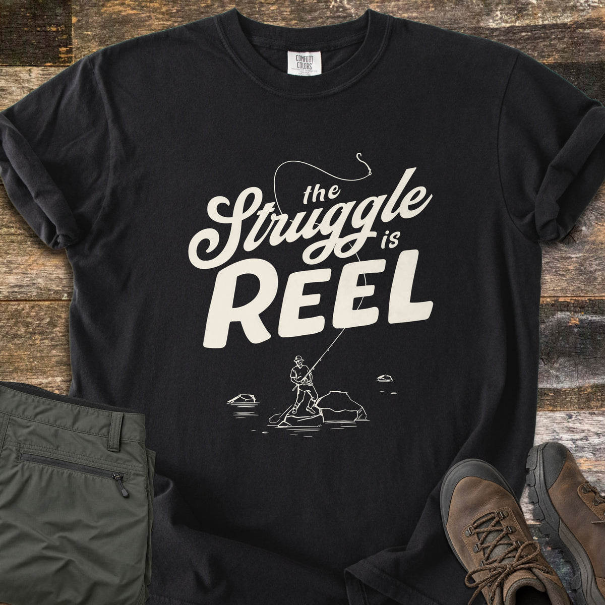 The Struggle Is Reel Comfort Colors T-shirt in Black, laid flat. Cream text reads the Struggle is REEL under an arcing fishing line with a hook, above a sketch of an angler casting from a rock in water. Folded olive pants and brown hiking boots sit on wood planks.