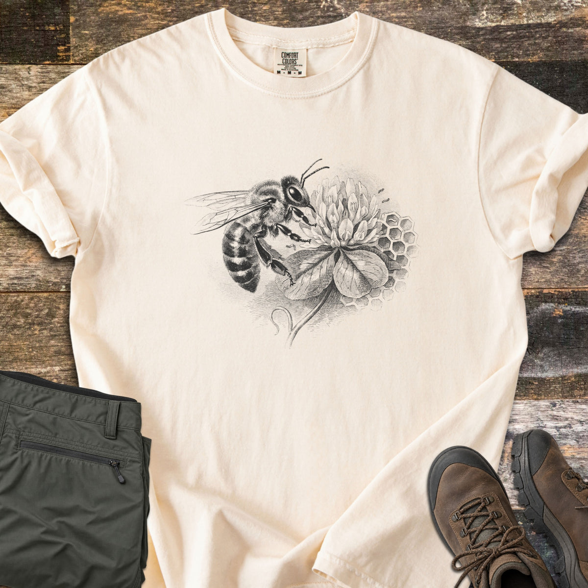 Honeybee On Clover Comfort Colors T-shirt in Ivory, laid flat. A detailed black engraving-style illustration of a honeybee on a clover blossom with honeycomb cells behind it, no words. Sleeves rolled, on weathered wood planks beside olive hiking pants and brown hiking boots.