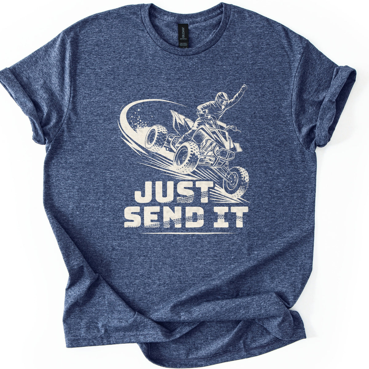 ATV Just Send It Comfort Colors T-shirt in Heather Navy, laid flat. A cream illustration of a helmeted rider on an airborne quad ATV, one fist raised, kicking up dust above the words Just Send It in bold tire-tread letters. The shirt lies on a white surface with cuffed sleeves.