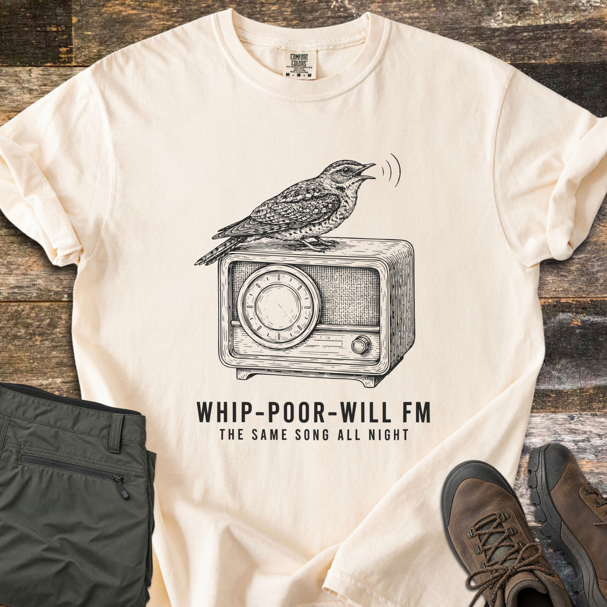 Whip-Poor-Will FM Comfort Colors T-shirt in Ivory, laid flat. A sketched whip-poor-will sings atop a vintage radio above the words Whip-Poor-Will FM, The Same Song All Night. It lies with rolled sleeves on weathered wood beside folded olive pants and brown hiking boots.