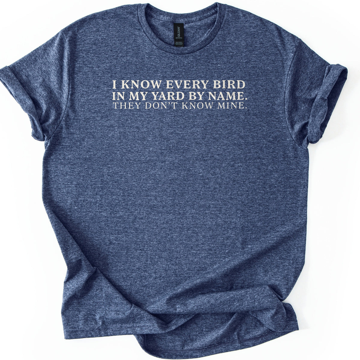 I Know Every Bird in My Yard by Name Comfort Colors T-shirt in Heather Navy, laid flat. Cream serif text across the chest reads I KNOW EVERY BIRD IN MY YARD BY NAME. THEY DON'T KNOW MINE. The sleeves are cuffed and the shirt lies on a plain white background.