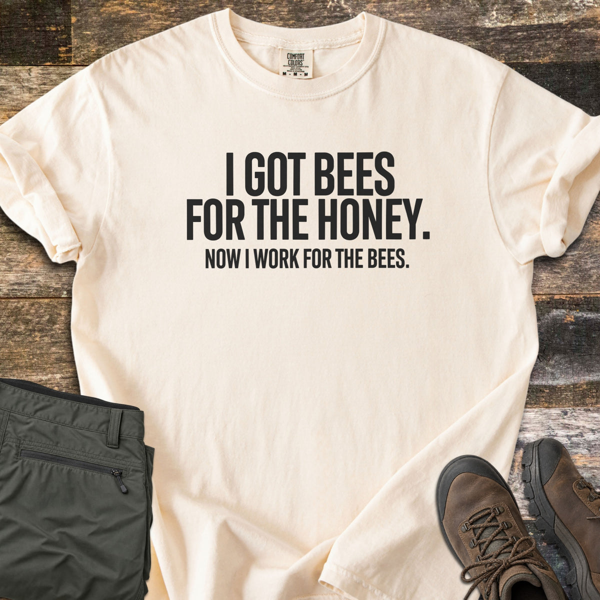 Work For The Bees Comfort Colors T-shirt in Ivory, laid flat. Black block letters read I GOT BEES FOR THE HONEY. NOW I WORK FOR THE BEES. The shirt lies with cuffed sleeves on weathered wood planks beside folded olive green pants and brown hiking boots.