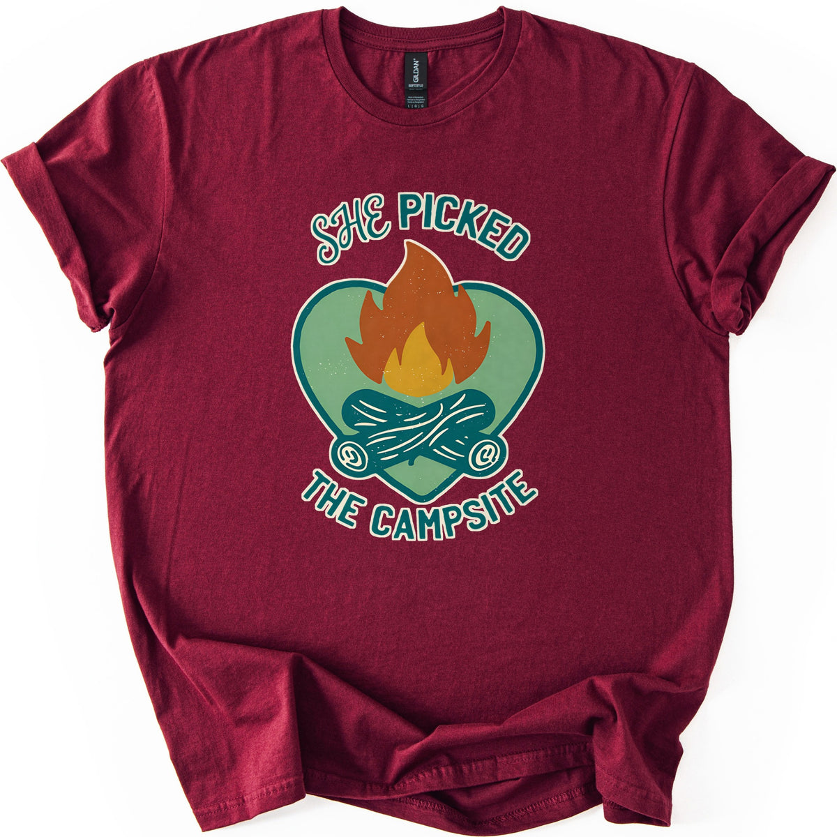 She Picked The Campsite Comfort Colors T-shirt in Maroon, laid flat. An orange and yellow campfire on crossed teal logs inside a sage green heart, with the words SHE PICKED above and THE CAMPSITE below. The shirt lies on a white surface with its sleeves cuffed.