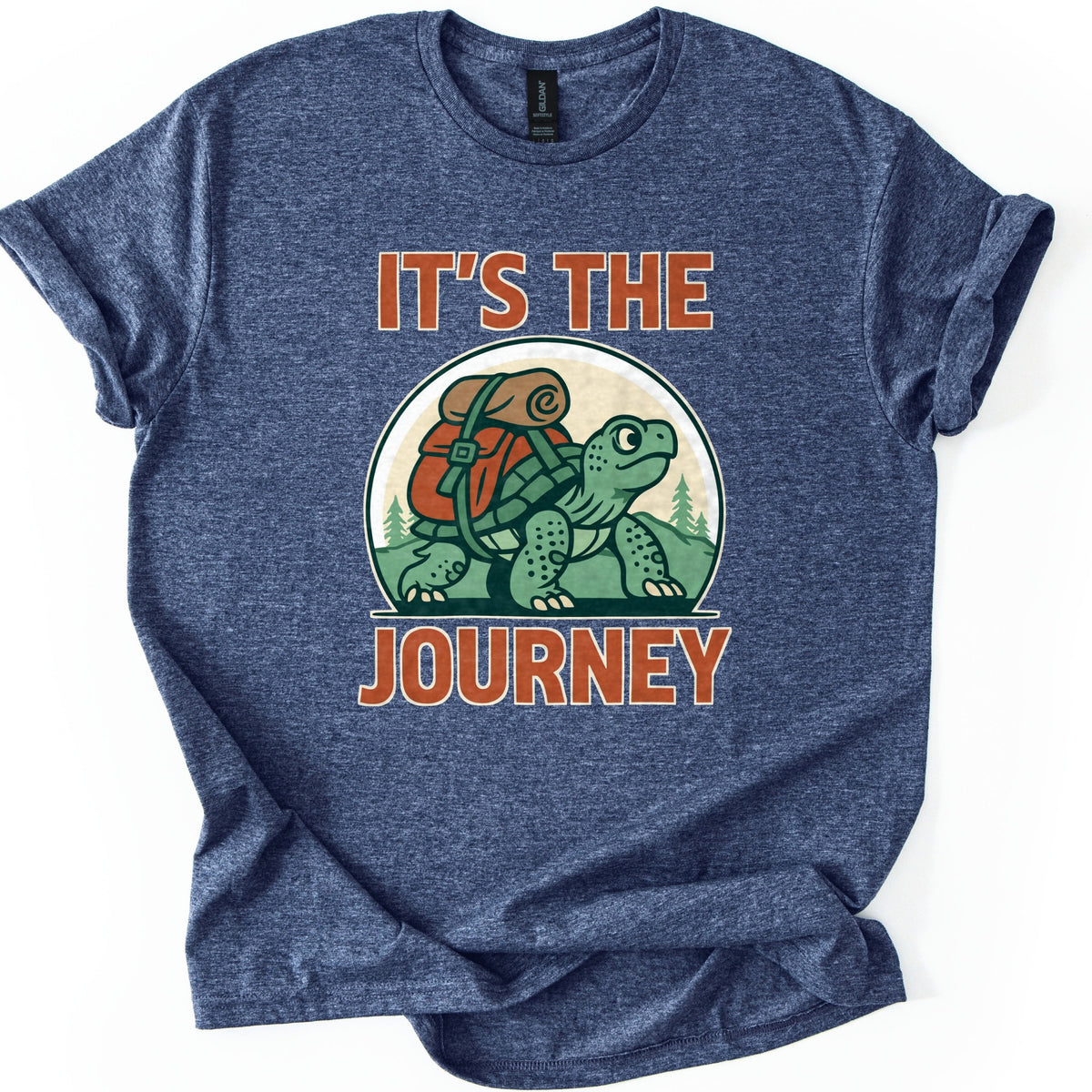 Slow Turtle Hiker Journey Comfort Colors T-shirt in Heather Navy, laid flat. A green cartoon turtle carrying a red backpack with a rolled sleeping bag walks past pine trees, framed by the words It's the Journey in rust-orange letters. Sleeves are cuffed on a white background.