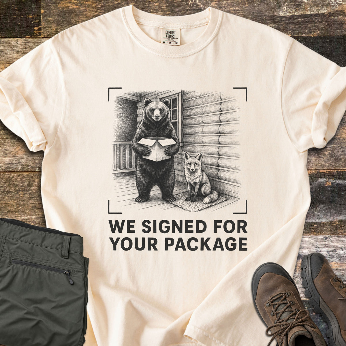 We Signed For Your Package Comfort Colors T-shirt in Ivory, laid flat. A black-ink sketch shows a bear holding an open box beside a seated fox on a log cabin porch, framed by camera corner marks above the words We Signed For Your Package. Olive pants and brown hiking boots sit on wood planks.
