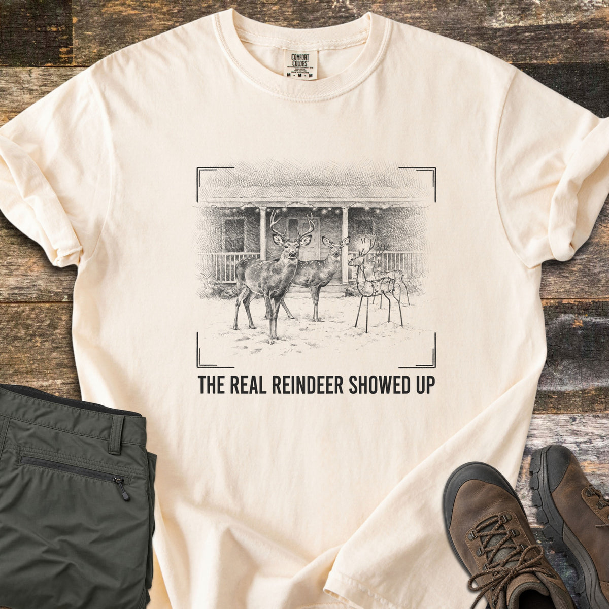The Real Reindeer Showed Up Comfort Colors T-shirt in Ivory, laid flat. A sketch of a buck and doe standing in snow next to a wire reindeer lawn decoration before a porch, inside camera viewfinder corners, above the words The Real Reindeer Showed Up. Olive pants and brown boots on rustic wood.