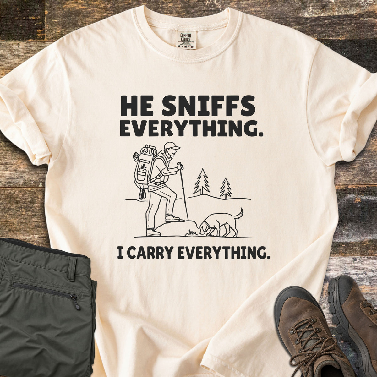 He Sniffs Everything. I Carry Everything Comfort Colors T-shirt in Ivory, laid flat. Black line art of a hiker with a large backpack and trekking pole beside a dog sniffing a rock, between the words He Sniffs Everything. and I Carry Everything. Olive pants and brown hiking boots sit on weathered wood.