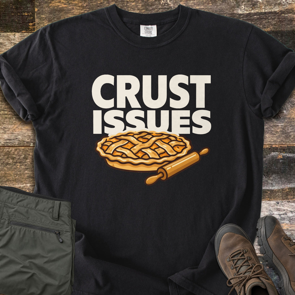 Crust Issues Comfort Colors T-shirt in Black, laid flat. The words CRUST ISSUES in off-white block letters sit above a cartoon lattice-top pie and a wooden rolling pin. Olive pants and brown hiking boots lie beside it on a rustic wood surface.