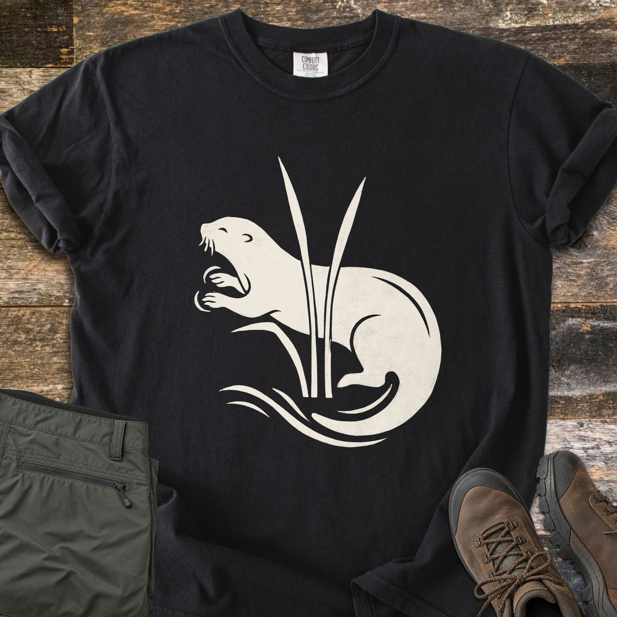Otter In The Reeds Comfort Colors T-shirt in Black, laid flat. A cream silhouette of an otter with open mouth and raised paws curls behind two tall reeds above wavy water lines, no words. Folded olive hiking pants and brown hiking boots lie on weathered wood.